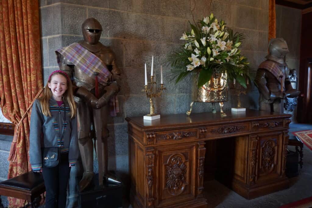 Dromoland Castle - girl wearing dark shirt and pants stands beside lobby desk with flowers and candles flanked by two suits of armour draped in a lavender tartan.