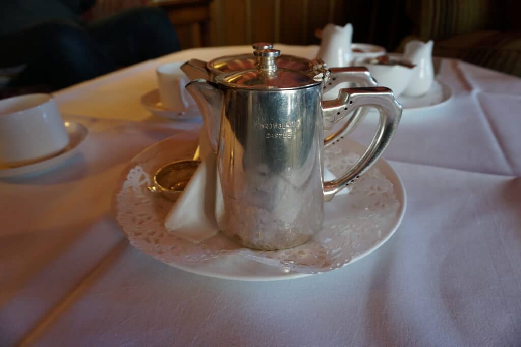 Silver tea pot with words Dromoland Castle engraved on it sitting on white saucer with doilie. White cups, saucers, cream and sugar in background on white tablecloth.
