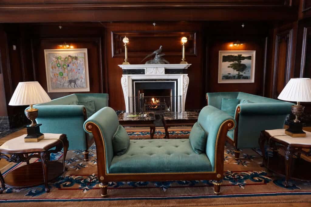 Seating area in Powerscourt Hotel lobby in front of fireplace with white mantel, two teal coloured sofas facing each other and a settee facing the fireplace, two ornate side tables with lamps.