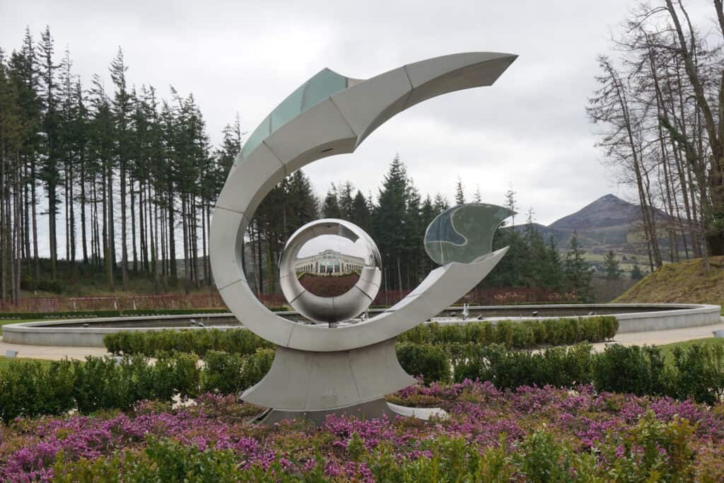 Sculpture in Powerscourt Gardens with hotel reflected in circular orb, purple flowers and green shrubbery at base and Wicklow Mountains in the distance.