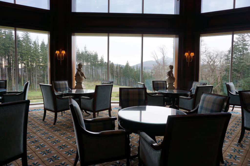 Sugar Loaf lounge at the Powerscourt Hotel - round tables and chairs in room with large glass windows facing trees and mountains in the distance.