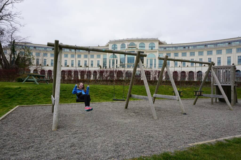 Girl in blue coat, black pants and pink shoes on wooden swing set on playground with the Powerscourt Hotel in background.