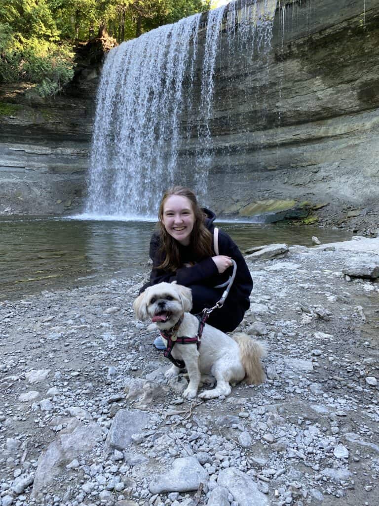 Young woman in dark clothes kneeling with light coloured dog in front of Bridal Veil Falls, Kagawong, Manitoulin Island.