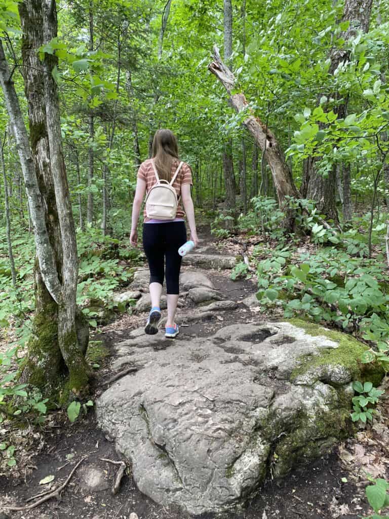 Young woman wearing small backback and carrying water bottle hiking over large rocks through woods.