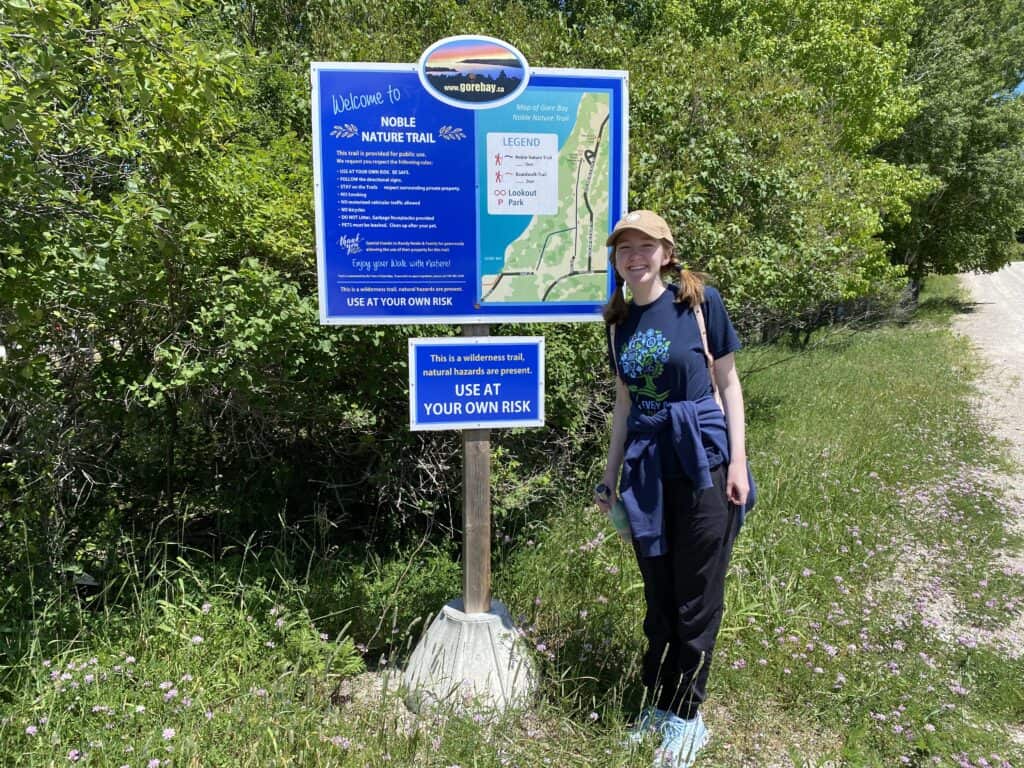 Young woman in dark pants and t-shirt wearing ballcap stands at entrance sign for Noble Nature Trail in Gore Bay on Manitoulin Island, Ontario, Canada.