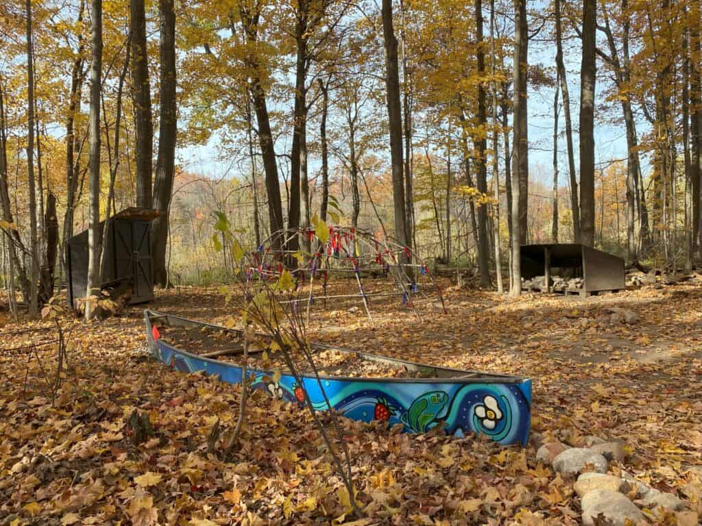 Wooded area with leaves on ground and colourful painted canoe with indigenous symbols in front of structure for sweat lodge.