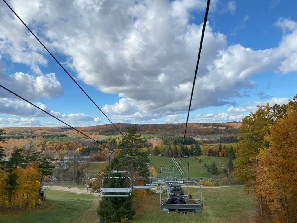 Chairlifts going down ski hill at Kelso Conservation Area with colourful fall trees stretching into the distance and blue sky with large fluffy white clouds.