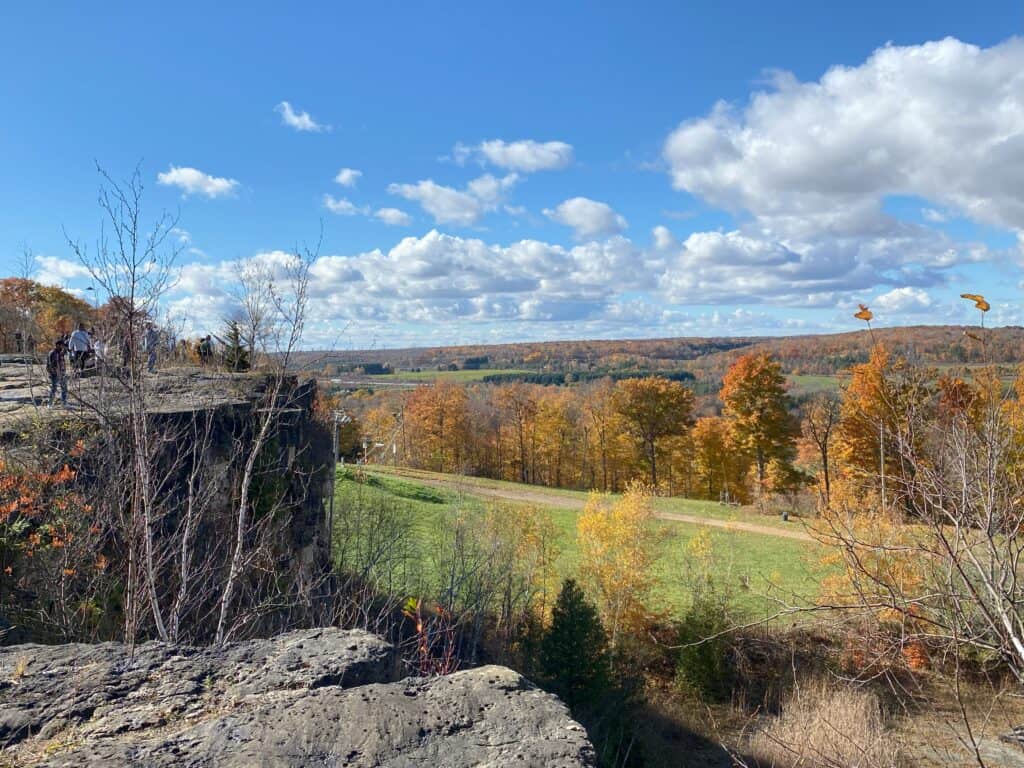 On a beautiful fall day with blue sky and fluffy white clouds, people standing on rocky escarpment looking at view of green ski hill and orange and yellow trees stretching into the distance.