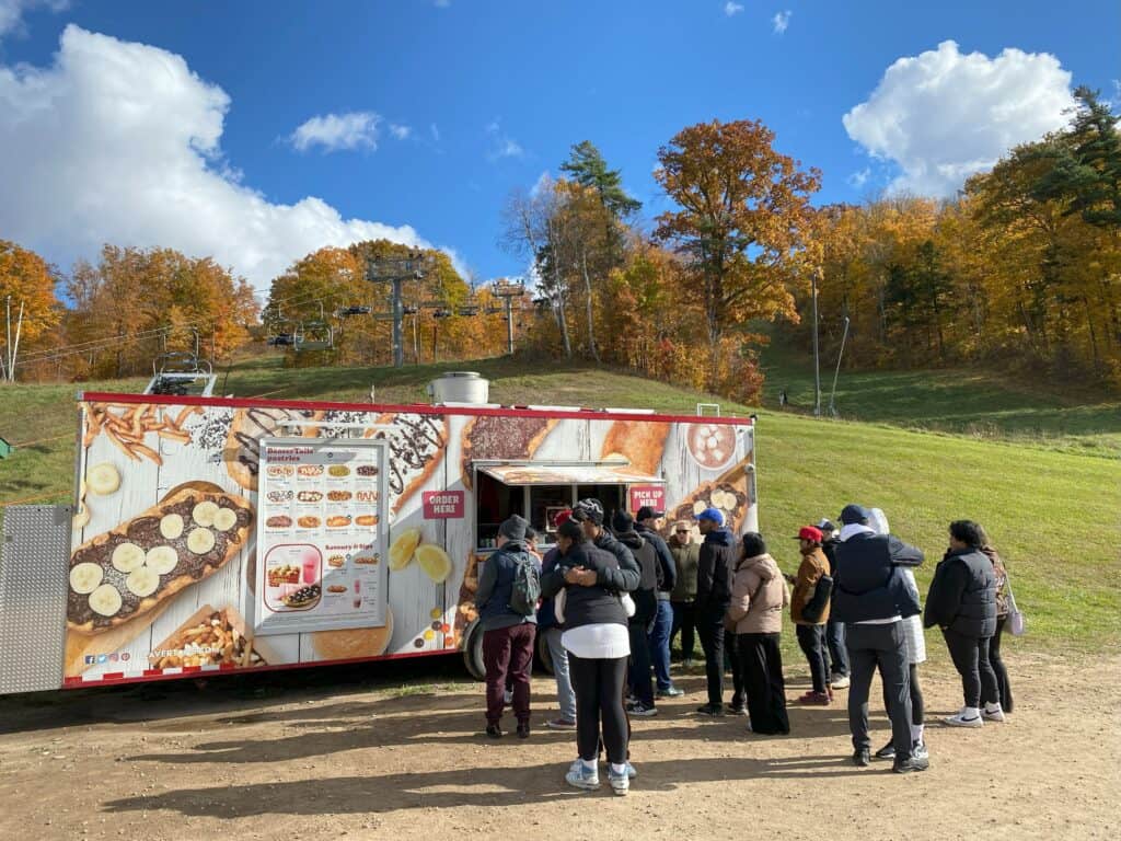 Group of people lined up at BeaverTails food truck at Kelso Conservation Area with ski lift and fall trees in background.