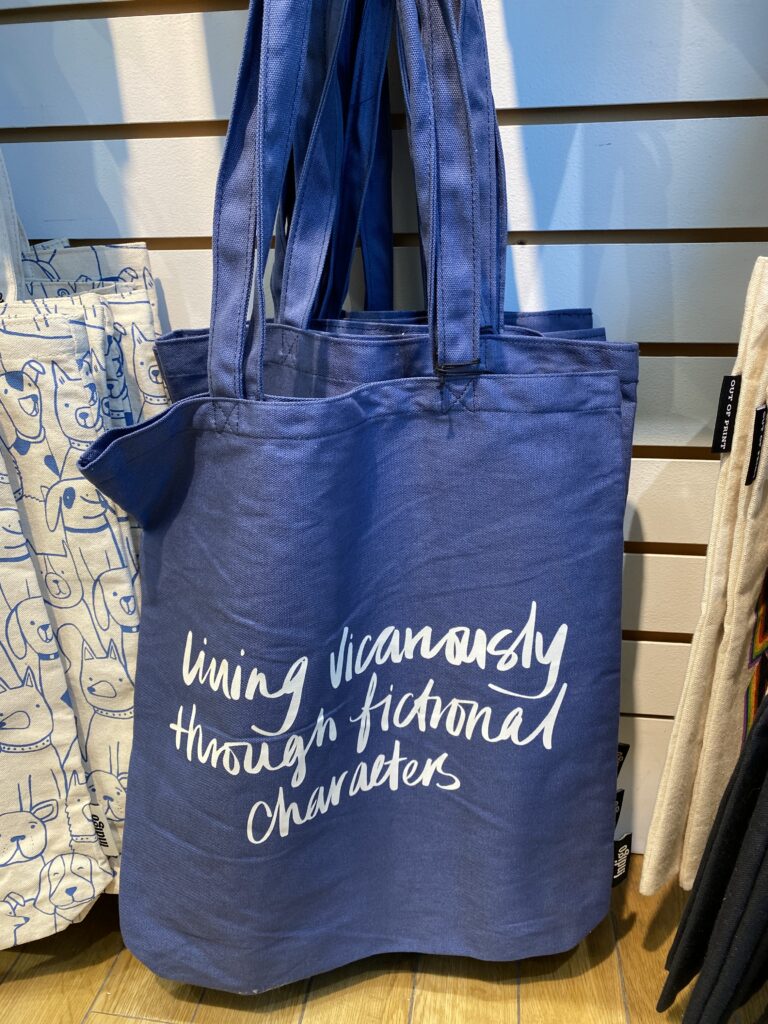 Blue tote bag with cursive white writing that reads "living vicariously through fictional characters".