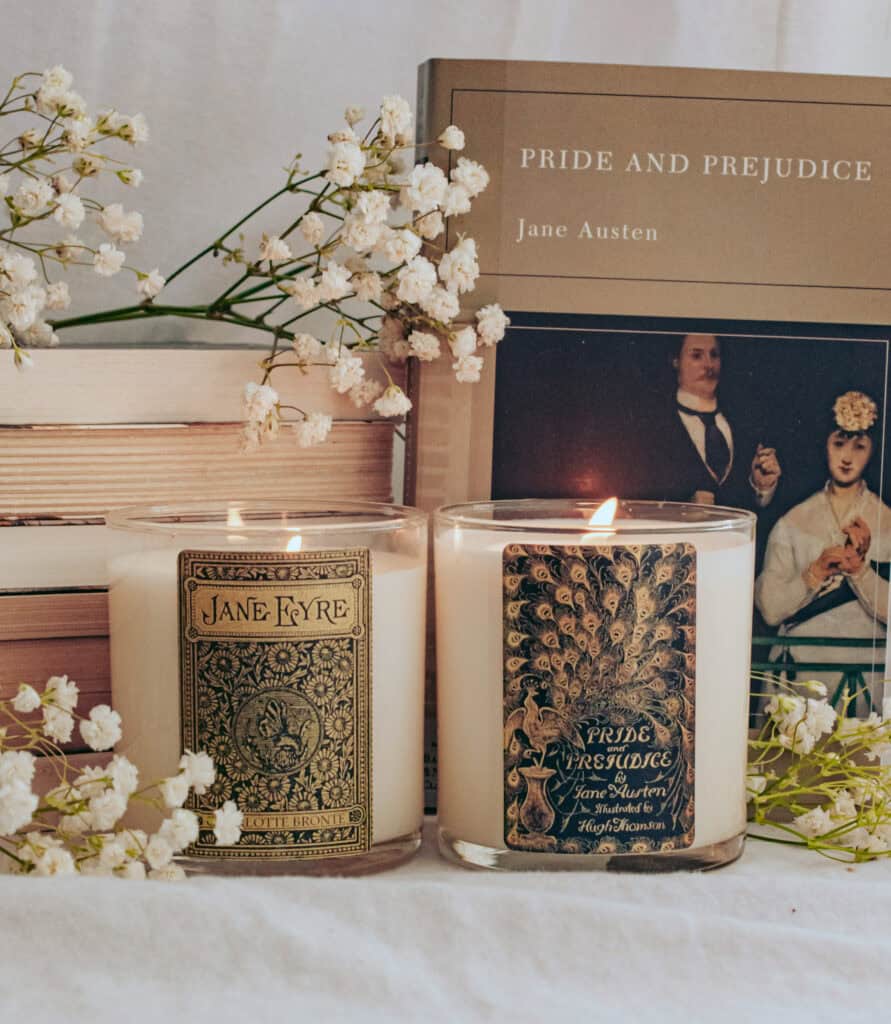 Jane Eyre and Pride and Prejudice cream coloured candles with black and gold label sit side by side with stack of books, baby's breath flowers and cover of Pride and Prejudice book in background.