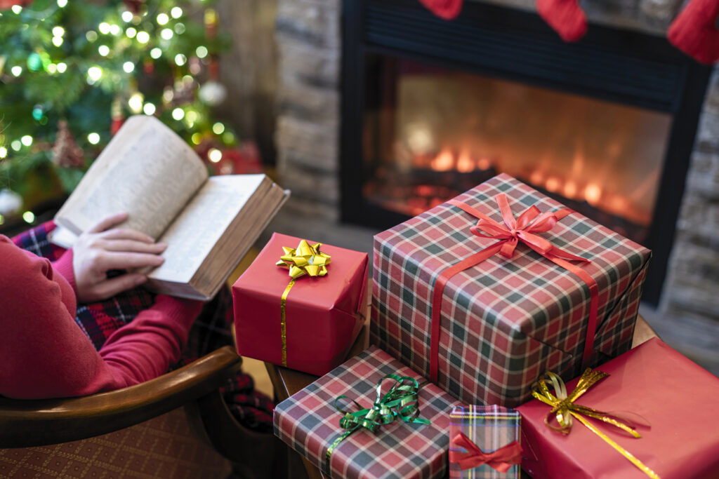 Partially visible woman in red shirt and red plaid pants  reading in front of fireplace beside stack of wrapped gifts with lit Christmas tree visible in background.