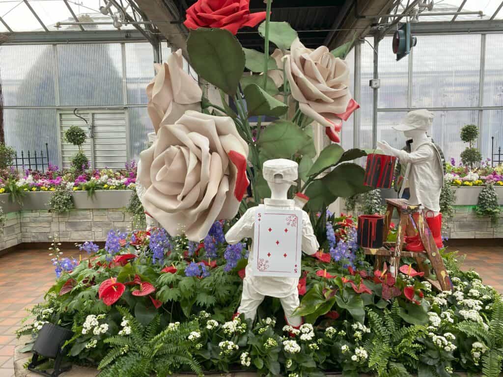 Breezeway display with two painters dressed in white clothes with red boots and playing cards on backs standing in greenery and flowers painting large white roses red - Alice in Bloomland at Royal Botanical Gardens.