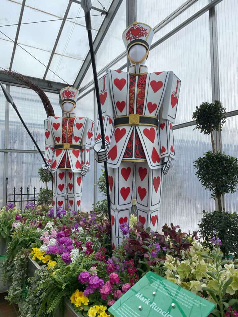 Two soldiers made from hearts playing cards holding swords standing amidst flowers and greenery.