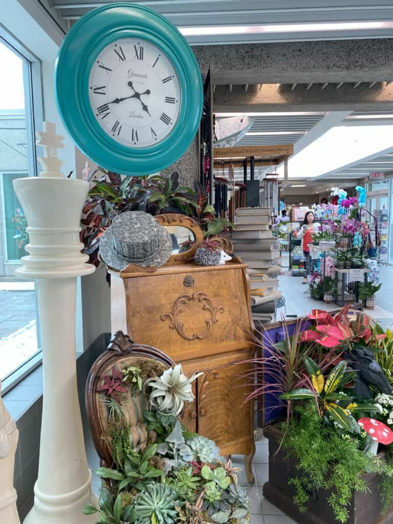 Whimsical display of flowers, furniture and odds and ends with blue hanging clock - Royal Botanical Gardens Alice in Bloomland showcase.