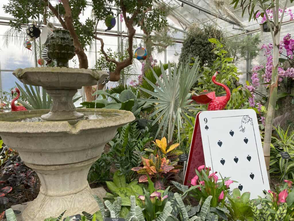 Mediterranean Garden stone water fountain surrounded by greenery, flowers, ceramic flamingoes, colourful teapots hanging from branches and a large 9 of spades playing card.