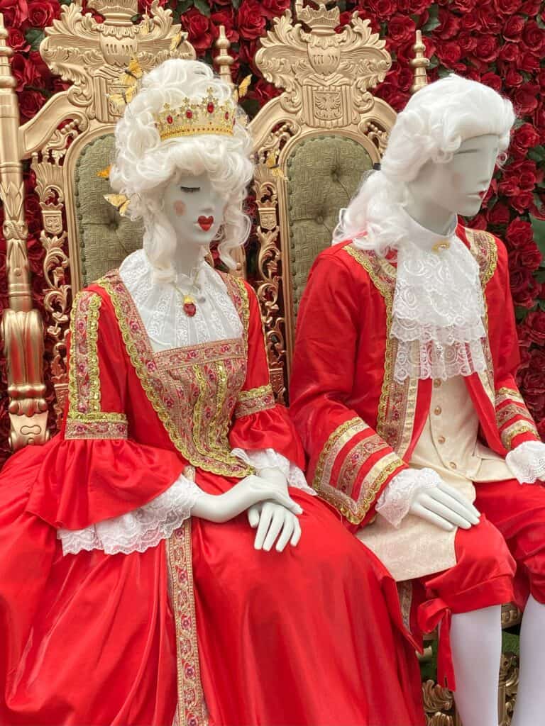 Male and female mannequins wearing red, gold and white costumes with white wigs sit on gold chairs in front of display of red roses at RBG Alice in Bloomland.