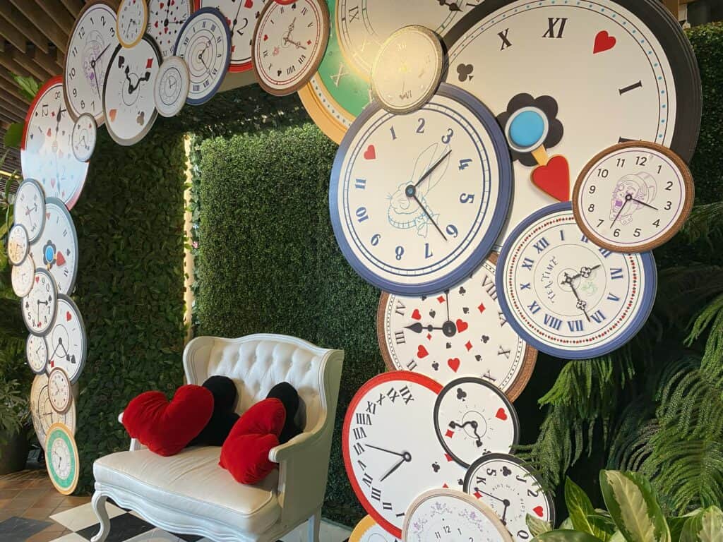 White loveseat with two red and two black heart-shaped cushions in front of greenery surrounded by arch display  of clocks with Alice in Wonderland theme - Alice in Bloomland at Royal Botanical Gardens.